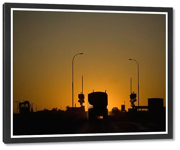 A freight truck and car stopped at a railroad crossing silhouetted against the setting sun