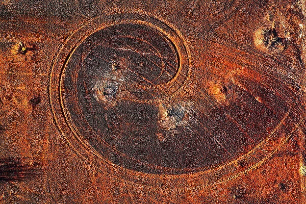 Abstract swirl pattern from car tyre tracks in red dirt, illuminated by dusk sunlight, remote rural Australia
