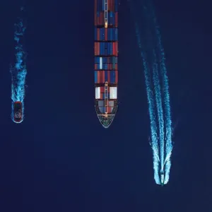 Vue aérienne d'un navire cargo guidé par un remorqueur et une barque de pilotage dans le port.
