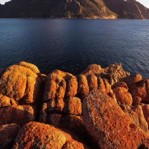 Sunrise at Wineglass Bay lights up lichen covered boulders with the Hazards mountain range beyond, Freycinet National Park, Tasmania, Australia