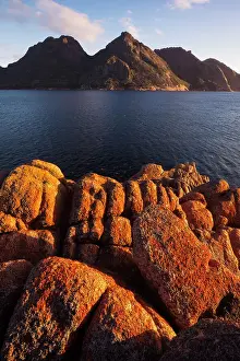 Sunrise at Wineglass Bay lights up lichen covered boulders with the Hazards mountain range beyond, Freycinet National Park, Tasmania, Australia